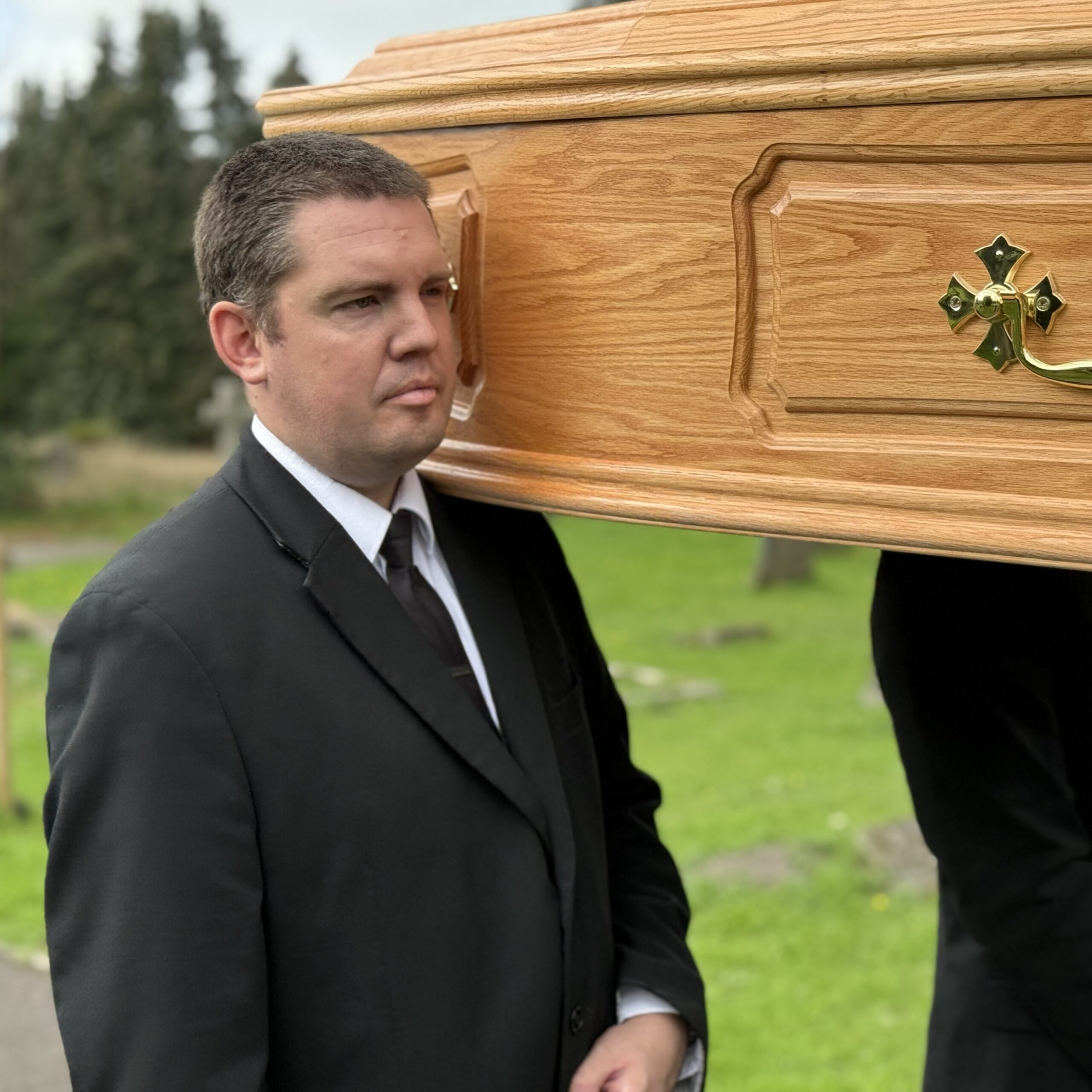 Man in black suit, carriers wooden coffin on shoulder.
