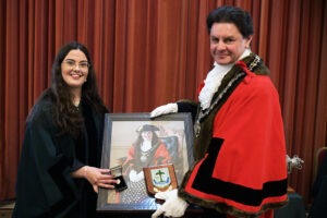 Photograph of Councillor Kathryn Macdermid, wearing a green Council robe, and Mayor of Chippenham, Councillor Matthew Bragg, wearing the Mayoral robe. They are holding a framed portrait of Councillor Macdermid wearing the Mayoral Robe and an award plaque, set against a red curtain backdrop. Photograph of Councillor Kathryn Macdermid, wearing a green Council robe, and Mayor of Chippenham, Councillor Matthew Bragg, wearing the Mayoral robe. They are holding a framed portrait of Councillor Macdermid wearing the Mayoral Robe and an award plaque, set against a red curtain backdrop.