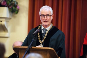 Photograph of Councillor Nick Murry, wearing a green Council robe and a large gold chain standing behind a wooden podium with a microphone. Photograph of Councillor Nick Murry, wearing a green Council robe and a large gold chain standing behind a wooden podium with a microphone.