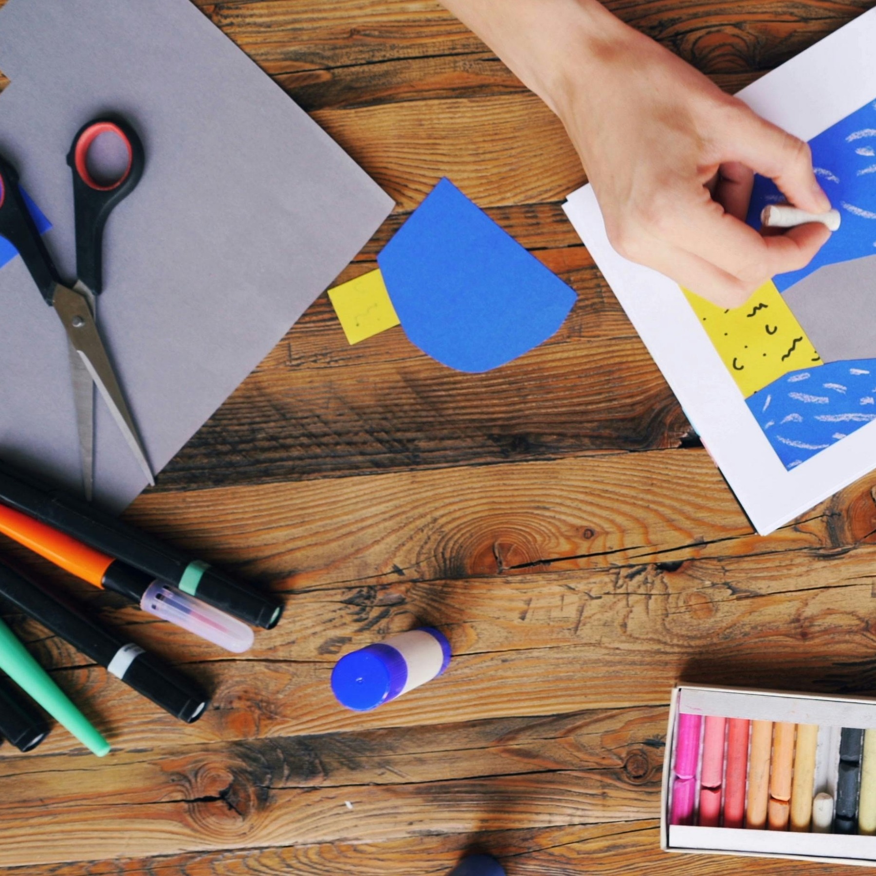 A wooden table with someone drawing using blue and yellow crayons. Grey, blue & yellow paper, scissors, glue, and colouring pens scatter across the table.