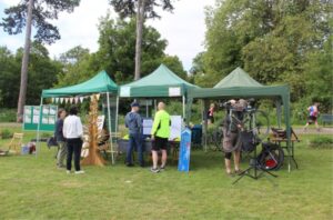 Participants engage at green tents during an outdoor event, with trees and runners in the background. Participants engage at green tents during an outdoor event, with trees and runners in the background.
