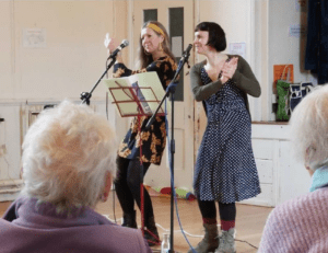 Photograph of two women performing in front of an audience, with one singing into a microphone and the other clapping. The setting is a community or recreational room, featuring microphones on stands, a music stand with sheet music, and elderly audience members seated in the foreground. Photograph of two women performing in front of an audience, with one singing into a microphone and the other clapping. The setting is a community or recreational room, featuring microphones on stands, a music stand with sheet music, and elderly audience members seated in the foreground.