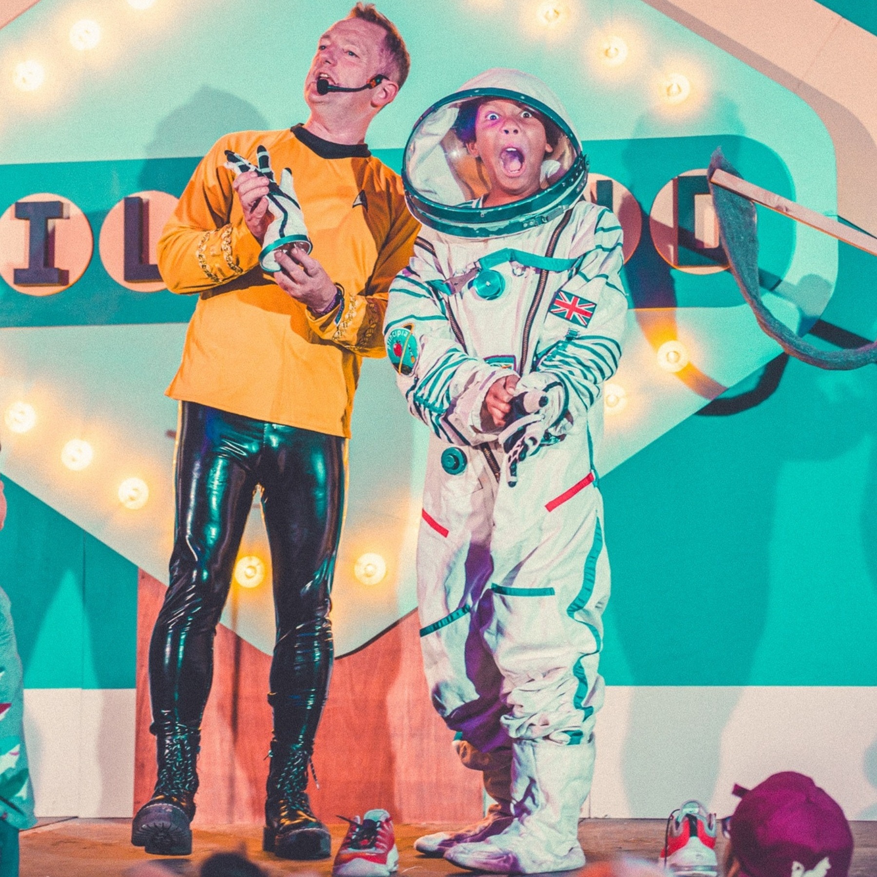 Adult in a yellow shirt and black trousers, wearing a microphone and holding a model rocket, alongside a child in a spacesuit.