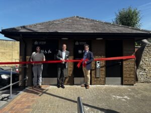 Photograph of Matt Kirby, Deputy Chief Executive, Will Tidmarsh, Head of Environmental Services, and Mayor of Chippenham, Cllr Matthew Bragg, standing in front of a newly refurbished toilets in Borough Parade Car Park, holding a large red ribbon for a ribbon-cutting ceremony. Building features brown wooden siding, black doors with accessibility and restroom signs, and a clear blue sky overhead. Photograph of Matt Kirby, Deputy Chief Executive, Will Tidmarsh, Head of Environmental Services, and Mayor of Chippenham, Cllr Matthew Bragg, standing in front of a newly refurbished toilets in Borough Parade Car Park, holding a large red ribbon for a ribbon-cutting ceremony. Building features brown wooden siding, black doors with accessibility and restroom signs, and a clear blue sky overhead.