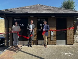 Photograph of Matt Kirby, Deputy Chief Executive, Will Tidmarsh, Head of Environmental Services, and Mayor of Chippenham, Cllr Matthew Bragg, standing in front of a newly refurbished toilets in Borough Parade Car Park, cutting a large red ribbon for a ribbon-cutting ceremony. Building features brown wooden siding, black doors with accessibility and restroom signs, and a clear blue sky overhead. Photograph of Matt Kirby, Deputy Chief Executive, Will Tidmarsh, Head of Environmental Services, and Mayor of Chippenham, Cllr Matthew Bragg, standing in front of a newly refurbished toilets in Borough Parade Car Park, cutting a large red ribbon for a ribbon-cutting ceremony. Building features brown wooden siding, black doors with accessibility and restroom signs, and a clear blue sky overhead.