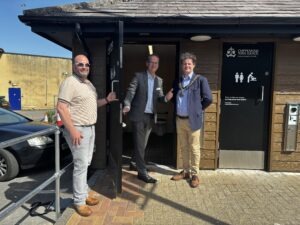 Photograph of Matt Kirby, Deputy Chief Executive, Will Tidmarsh, Head of Environmental Services, and Mayor of Chippenham, Cllr Matthew Bragg, standing in front of a newly refurbished toilets in Borough Parade Car Park. Building features brown wooden siding, black doors with accessibility and restroom signs, and a clear blue sky overhead. Photograph of Matt Kirby, Deputy Chief Executive, Will Tidmarsh, Head of Environmental Services, and Mayor of Chippenham, Cllr Matthew Bragg, standing in front of a newly refurbished toilets in Borough Parade Car Park. Building features brown wooden siding, black doors with accessibility and restroom signs, and a clear blue sky overhead.