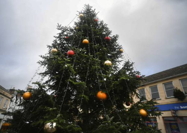 Christmas tree Photograph of a large outdoor Christmas tree decorated with red, gold, and orange baubles and string lights. Tree is set against a cloudy sky with surrounding buildings visible in the background.
