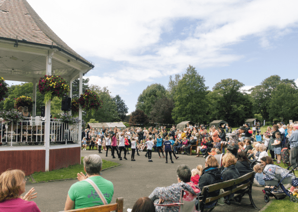 Summer event John Coles Park Photograph of an outdoor summer event at John Coles Park featuring a group of children performing a dance in front of a seated audience near the band stand adorned with hanging flower baskets. The scene includes a crowd of spectators, lush green trees, and a partly cloudy sky, highlighting a lively and family-friendly atmosphere.