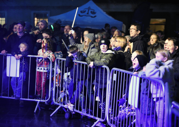 Christmas crowd Photograph of a crowd of people, including children and adults, standing behind metal barricades at a Christmas Light Switch On event at night. Blue and purple lighting highlights faces, with some individuals wearing winter clothing and a tent visible in the background.