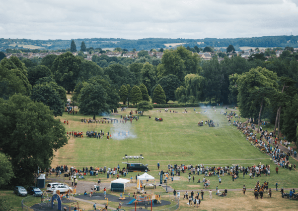 Summer event ECWS Photograph of a Monkton Park during the Battle of Chippenham re-enactment event with numerous people gathered across a wide grassy field surrounded by dense trees. The scene includes a playground area, tents, parked cars, and groups of people scattered throughout, indicating a community gathering or festival on a cloudy day.