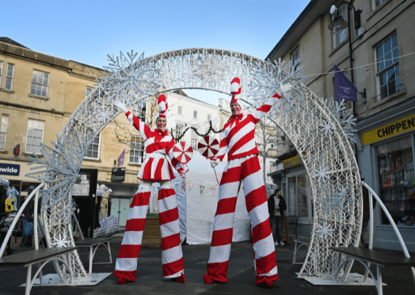 Christmas stilts Photograph of two performers on stilts dressed in red and white striped costumes resembling candy canes, standing under a decorative arch adorned with white snowflake lights. The festive street scene includes historic buildings and shops, highlighting a holiday or winter celebration event.