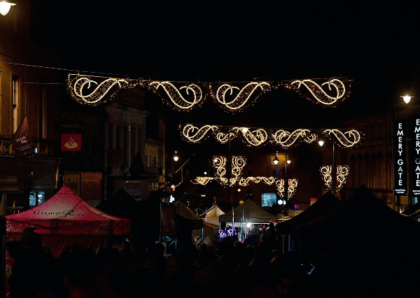 Christmas lights Photograph of a night time street market on Chippenham Highstreet decorated with illuminated festive light garlands hanging above. The scene features dark tents and stalls with people silhouetted against bright lights, highlighting a lively event atmosphere.