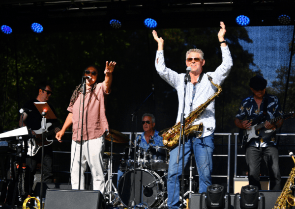Summer event stage Photograph of a live band performing on an outdoor stage with five musicians playing instruments including keyboard, drums, saxophone, and guitar. The scene features colorful stage lights, musicians wearing casual clothing, and an energetic atmosphere with one vocalist singing and another musician raising hands.