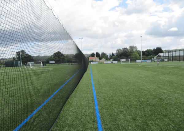 Stanley Park Photograph of an outdoor football pitch featuring bright blue boundary lines and a black net running diagonally across the foreground. The scene includes goalposts, light poles, trees, and a partly cloudy sky, suggesting a well-maintained sports facility at Stanley Park Sports Ground on a clear day.