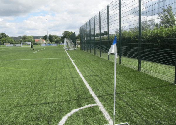 Stanley Park Photograph of an outdoor football pitch, a corner flag with blue and white colors, and a tall black fence along one side. The scene includes a goalpost in the distance and a person walking near the field under a partly cloudy sky.