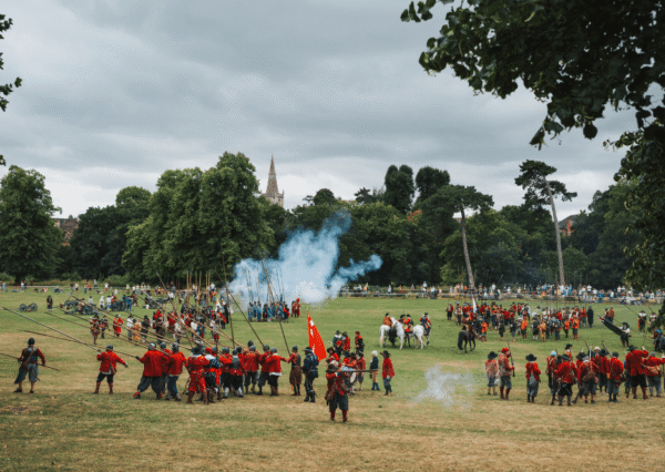 Summer event ECWS Photograph of a historical reenactment battle taking place in Monkton Park with participants dressed in period costumes, including soldiers in red uniforms holding pikes and cavalry on horseback. Smoke from gunfire is visible, with a backdrop of trees and a church steeple under a cloudy sky, capturing a dynamic and immersive scene.