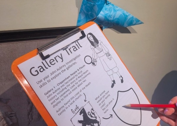 A printed black and white paper gallery trail on an orange clipboard, in front of a museum display of art.
