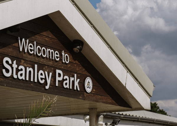 Stanley Park Photograph of a wooden and concrete entrance sign reading "Welcome to Stanley Park" with a small circular logo beneath the text. The sign is set against a partly cloudy sky, indicating an outdoor park entrance.