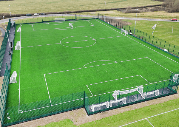 Stanley Park Aerial photograph of the Stanley Park Sports Ground 3G pitch with green turf, white boundary lines, and goalposts at each end. The field is enclosed by a tall green fence, with benches and a pathway surrounding it, situated near open grassy areas and a road with a few vehicles.