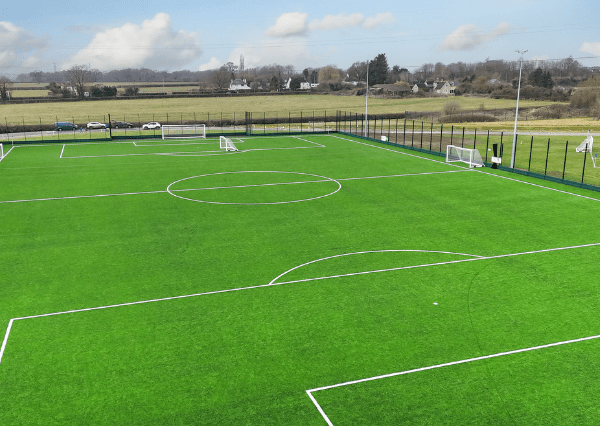 Stanley Park Aerial photograph of the Stanley Park Sports Ground 3G pitch with green turf, white boundary lines, and goalposts at each end. The field is enclosed by a tall green fence, with benches and a pathway surrounding it, situated near open grassy areas and a road with a few vehicles.