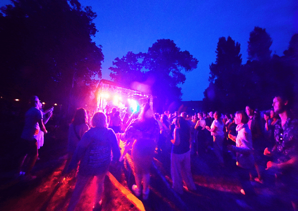 Summer event stage Photograph of an outdoor nighttime concert with a crowd gathered in front of a brightly lit stage emitting purple and orange lights. Attendees are standing and dancing on grass surrounded by trees under a dark blue sky.