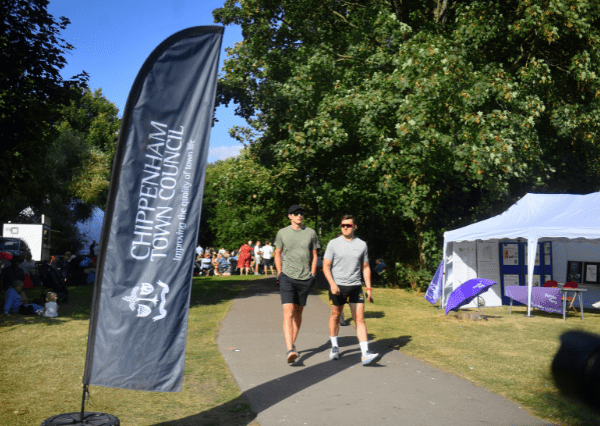 Summer event flags Photograph of an outdoor community event in a park featuring a tall black flag with "Chippenham Town Council" text and logo on the left side. Two casually dressed men walk on a paved path past a white tent with purple umbrellas, surrounded by green trees and other attendees in the background
