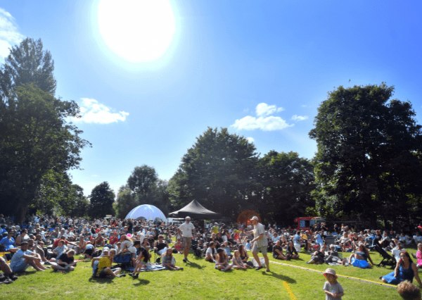 Summer event John Coles Park Photograph of a large outdoor gathering in a park on a sunny day with clear blue sky and bright sun overhead. Numerous people are sitting on grass, surrounded by tall trees, with tents and a food truck visible in the background, indicating a community event or festival.