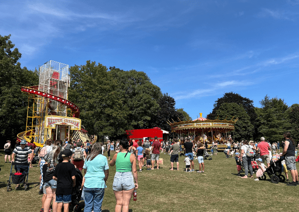 Summer event John Coles Park Photograph of an outdoor amusement park scene with families and children gathered around rides on a sunny day. Key elements include a tall, red and yellow helter-skelter slide on the left and a traditional carousel with horses on the right, set against a backdrop of green trees and blue sky.