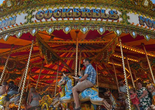 Summer event carousel Photograph of a colourful carousel with ornate decorations and bright lights, featuring a man riding a blue and yellow horse figure. Several other riders and onlookers are visible, highlighting a lively amusement park atmosphere during daytime.