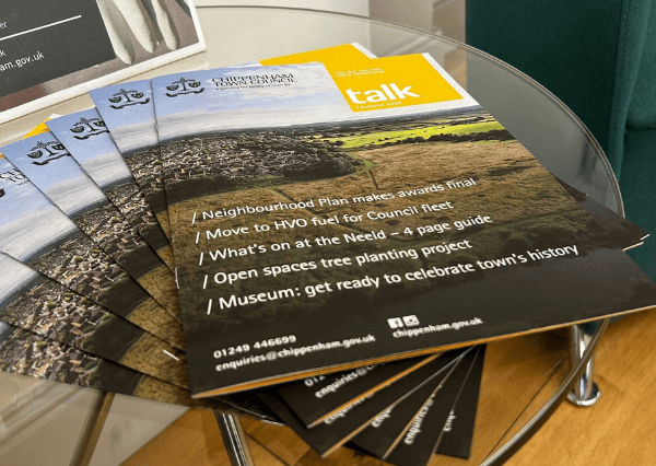 Talk image Photograph of several printed newsletters titled "Chippenham Town Council Talk" arranged in a fan shape on a glass table. The newsletters feature headlines about local initiatives such as Neighbourhood Plan awards, council fleet fuel transition, a Neeld What's On guide, tree planting projects, and a museum celebrating town history, with contact information and a yellow header.