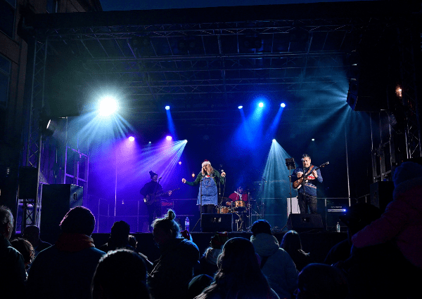 Christmas Stage Photograph of a live music concert featuring a band with a singer, guitarist, bassist, and drummer performing on a stage illuminated by blue, purple, and white spotlights. Audience silhouettes are visible in the foreground, highlighting an engaged crowd enjoying the Christmas Light Switch On.