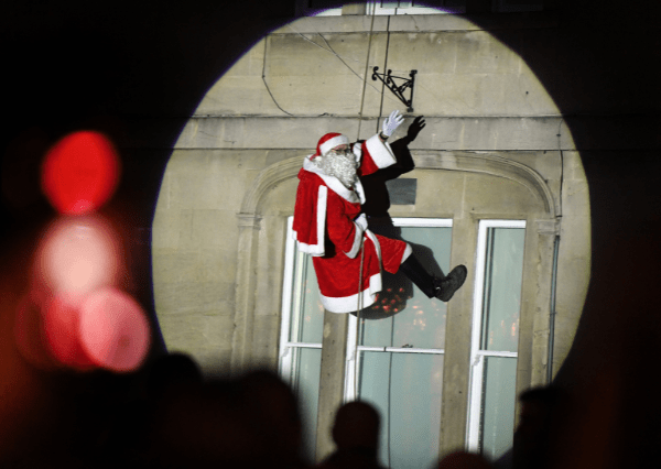 Christmas Santa Photograph of Santa Claus abseiling down a building at Christmas Light Switch on, illuminated by a circular spotlight. The scene includes a stone wall with windows and silhouettes of onlookers in the foreground, highlighting a festive or performance event.