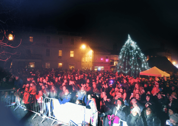 Christmas crowd 2 Photograph of a large crowd gathered outdoors at the Christmas Light Switch On, featuring a brightly lit Christmas tree and illuminated buildings in the background. The scene includes metal barricades in the foreground and people dressed in winter clothing, indicating a holiday celebration or public gathering.