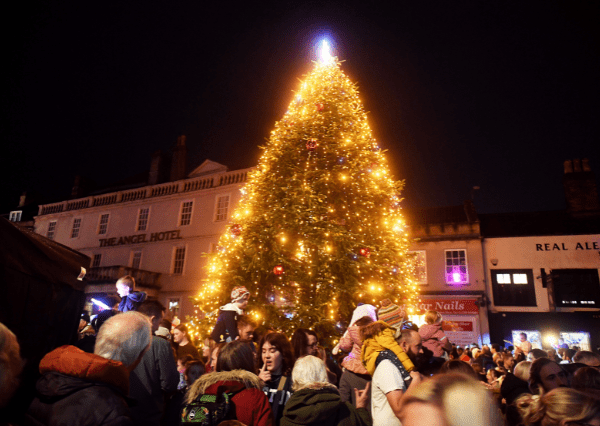 Christmas tree Photograph of a large, brightly lit Christmas tree in a town square at night, surrounded by a crowd of people celebrating. The tree is decorated with golden lights and red ornaments, with nearby buildings displaying shop signs.