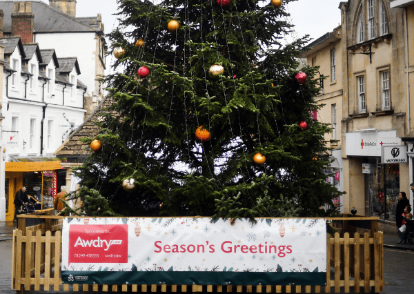 Christmas tree Photograph of a large outdoor Christmas tree decorated with red, gold, and orange ornaments, situated in a town square surrounded by buildings. A wooden fence encloses the tree, featuring a banner with "Season’s Greetings" and sponsor information from Awdry Law.