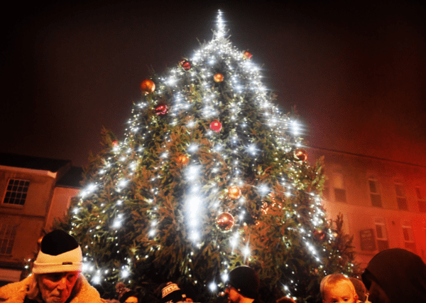 Christmas tree Photograph of a large outdoor Christmas tree decorated with white string lights and red ornaments, glowing brightly against a dark evening sky. Several people, including children wearing winter hats, are gathered around the tree, highlighting a festive holiday celebration in a town setting.