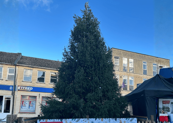 Christmas tree Photograph of a large, unlit Christmas tree placed in an outdoor town square surrounded by buildings and a black tent. The tree is enclosed by a festive white barrier with holiday-themed decorations, set against a clear blue sky.