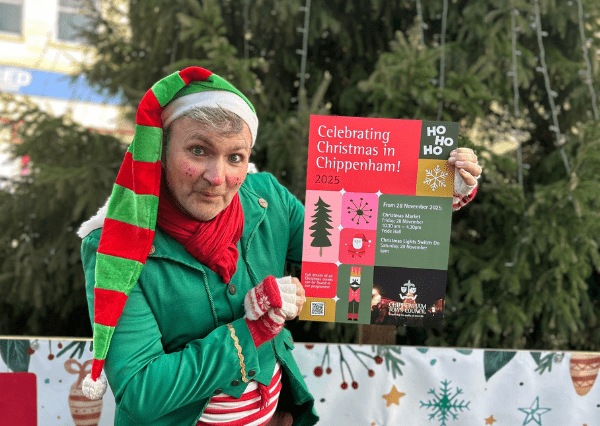 Christmas tree Photograph of Buddy McJingles holding a colourful flyer promoting Christmas celebrations in Chippenham for 2025. Flyer includes event details with icons of a Christmas tree, clock, Santa, and nutcracker, featuring red, green, black, and yellow sections with text about markets and festivities. Stood in front of the Christmas Tree.