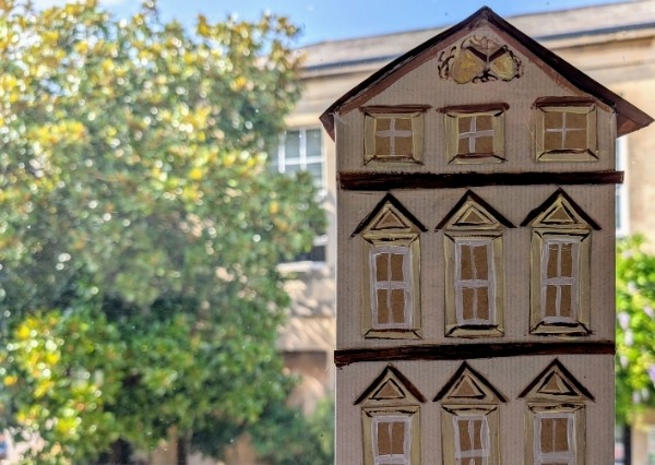 A 3D recycled cardboard house set against a background of trees.