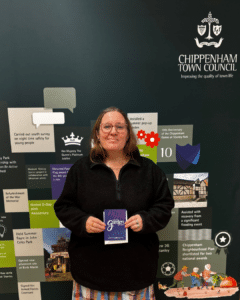 National Garden Voucher Photograph shows a woman holding a booklet titled “National Garden Gift Card” in front of a display celebrating Chippenham Town Council and community achievements. Background panels highlight initiatives and milestones including a 10th anniversary, volunteer projects, recreation facilities, heritage, sports, and national recognition, with council crest and speech-bubble accents.