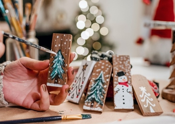 Chippenham Museum Festive Workshops 2026 An image showing a person painting snowy details on a small wooden Christmas tree ornament, with other hand-painted decorations nearby.
